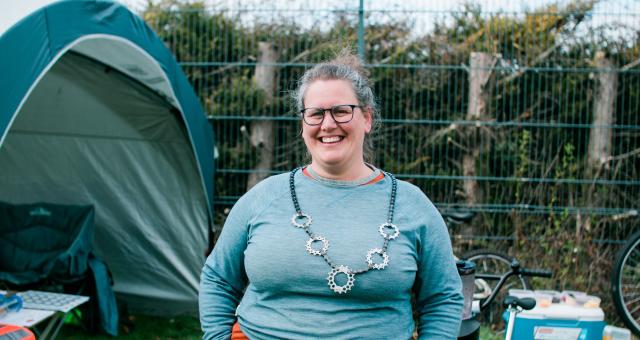 Emily Williams standing with her hands in her pockets, wearing a blue long-sleeved top, glasses and is smiling. She has a necklace made from a bike chain