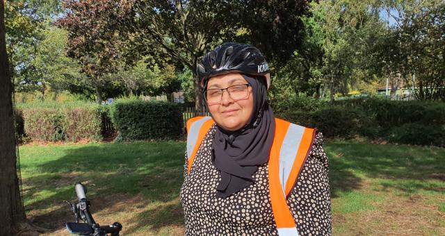 Fatima Patel is standing in front of a red bike wearing black rimmed glasses, an orange high vis jacket and a black head scarf underneath a cycling helmet