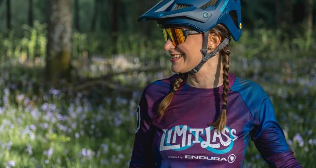 Fiona Finnie is standing in a sunny flower covered woodland wearing a cycle helmet, sunglasses and a t-shirt that reads Limitlass