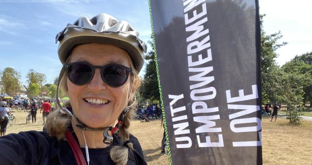 Gönül Ekmekçi Tekin is wearing sunglasses and a cycling helmet and is standing next to a black flag that reads ‘unity empowerment love’ there are crowds of people behind her some with cycles