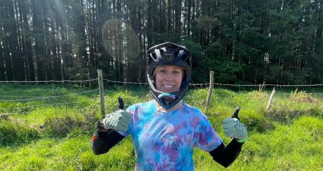 Georgina Tucker is standing on a mud track in front of a wire fence and wooded background with both thumbs up wearing a downhill cycling helmet