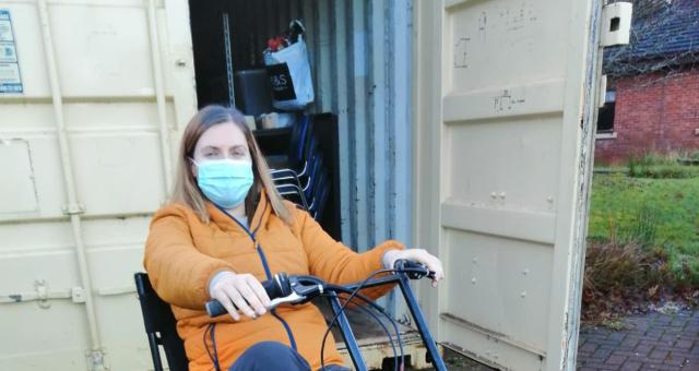Hannah Knox is sitting on a recumbent tricycle in front of a white shipping container she is wearing a blue face mask and yellow jacket