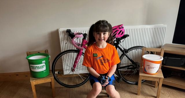 Heidi Barker is sitting on a chair in front of her pink bike. On either side of her bike are charity donation buckets and she is wearing an orange t-shirt and cycling shorts