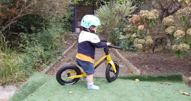 A little boy is riding the Hornit AIRO Balance Bike, a yellow balance bike. He's wearing a helmet and he's in a garden