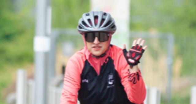 Humira Shahzad is cycling along a wide cycle path, wearing a red top, sunglasses and black cycle helmet. She is waving with one hand