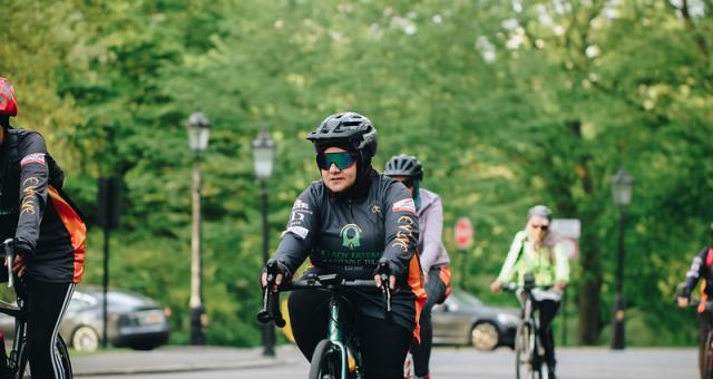 Iffat Tejani is cycling along a road with several other women. She is wearing sunglasses and a cycle helmet with a head scarf underneath