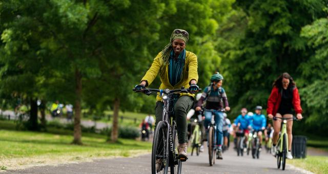 A large group of people is cycling along a paved path through green space, with trees behind them. There is a woman cycling on an e-bike at the front. She is wearing normal clothes and a headscarf. She is smiling