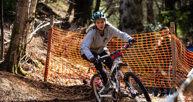 Jaz Morse is riding down a muddy woodland track on a mountain bike, there is an orange plastic fence and trees in the background