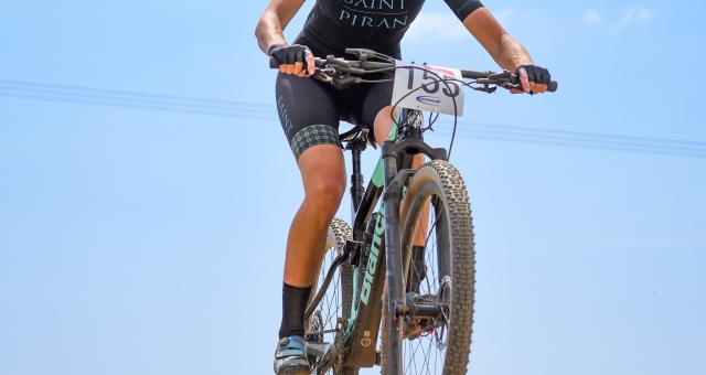Jenny Bolsom on her mountain bike wearing Saint Piran kit, a helmet and sunglasses. In the photo, her bike is up in the air above the gravel track below as she has just ridden over a bump.