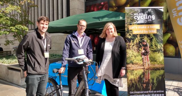 Three people (two men and a woman) are standing in front of a gazebo with a Cycling UK banner to the side. The man in the centre is holding an e-bike.