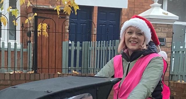 Katie Collier is riding a cargo bike in a pink high vis jacket and wearing a santa hat. At the front of the cargo bike there are children sitting under a black cover