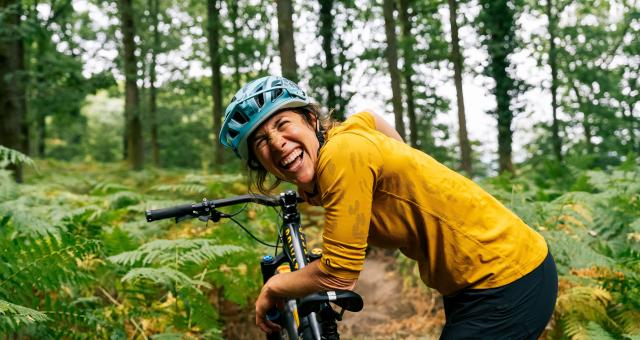 Katy Curd laughing while delivering mountain biking coaching in the Forest of Dean. She is leaning on the seat of a mountain bike. She is wearing a bright yellow top and cycling helmet.