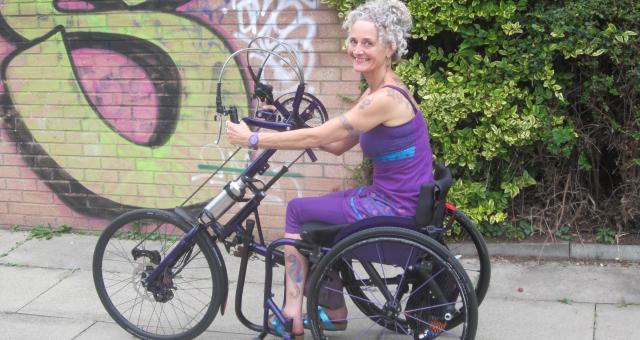 Kay Inckle is riding an adapted handcycle along a paved path. Behind her is a hedge and part of a brick wall with graffiti on it. She is wearing purple leggings and strappy top. She is smiling at the camera.