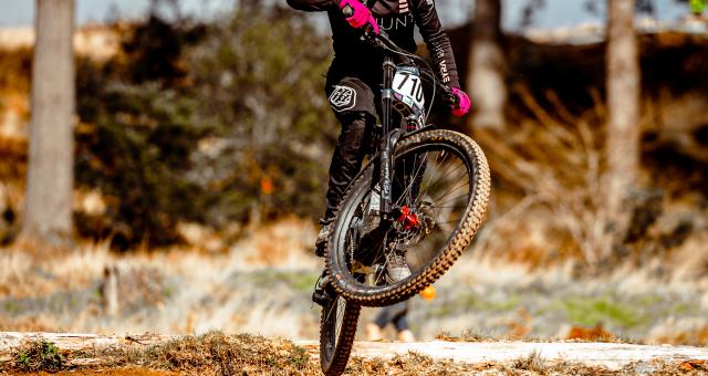 Kerry Wilson is performing a jump on a downhill off-road mountain bike track. She is wearing black mountain biking kit and a full face helmet. She has a race number (710) on her mountain bike handlebar.