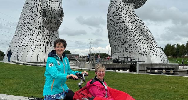 Kirsteen Ross pedals an adapted tricycle trishaw in front of the Kelpies in Falkirk. She is wearing a blue jacket and black leggings. She has a passenger in the trike, who has a bright blanket over her. Both are smiling at the camera.