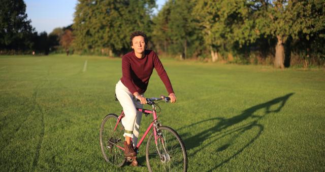 Laura Di Giacomo on her pink bike in the park in Oxford. She is wearing a red long-sleeved T-shirt, white trousers and brown boots. She has short, curly brown hair.