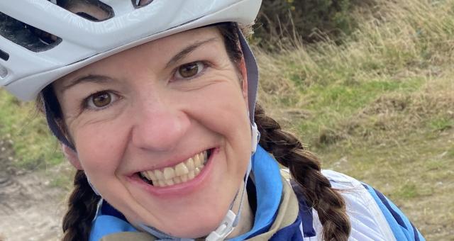 Lorna Breetzke shows off her blue and white Elgin Cycling Club kit. She has taken a selfie and is smiling at the camera. She has long brown hair in plaits and is wearing a cycle helmet. She’s out in the countryside and there’s a big gorse bush behind her.