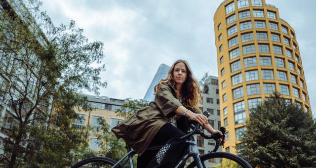 Lucy Poulden is sitting on a black road bike. She is on a road in front of a yellow tall building and is wearing a long green jacket and black trousers