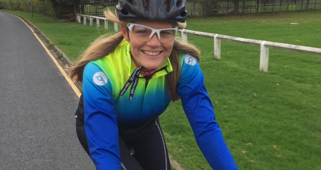 Lynn Bye rides her road bike togged up in Fat Lad at the Back cycling kit, a blue and yellow long-sleeved jersey and black leggings. She is wearing a helmet and cycling glasses; she has long blonde hair.