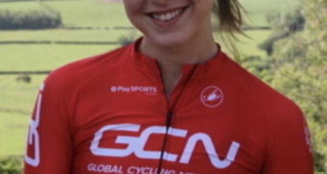 A portrait of cyclist Manon Lloyd in her red GCN cycling kit, with white GCN logo. She has long brown hair in a ponytail and is smiling at the camera.