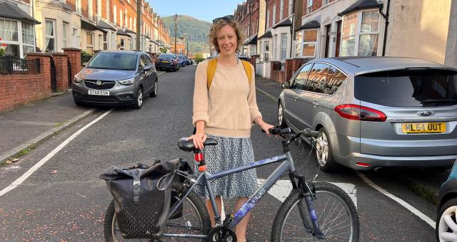 Meg Hoyt stands with her hybrid bike on a terraced street. She is wearing a yellow sweater and patterned skirt. She has a rucksack on and there are rear panniers on her bike.