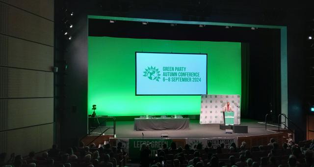 A man is standing on a stage to the right. A big green screen is displaying the Green Party logo and the words 'Green Party autumn conference 6-8 September 2024'