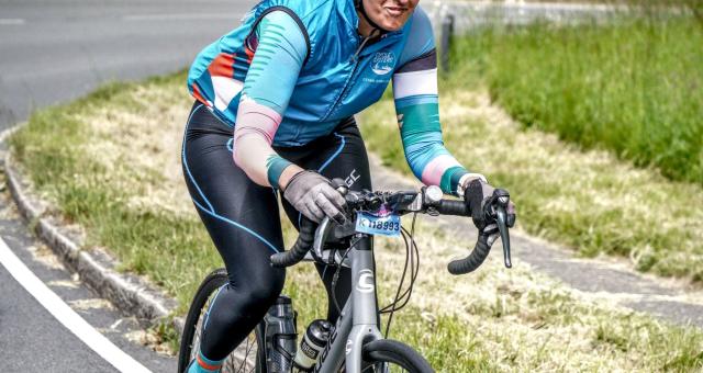 Nahida Hussain is riding a silver Cannondale road bike. She is wearing a Cycle Sisters cycling jersey and cycling leggings, as well as a helmet and cycling sunglasses. She has an event number fixed to her handlebar and is smiling.