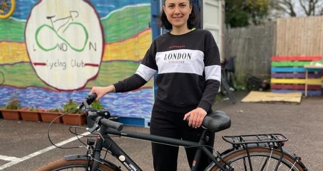 A woman is standing holding a bike. She is wearing black jeans and a black sweater with a grey stripe across it in which reads London Stratford. Behind her is an old shipping crate which has been painted with bright colours and a logo reading London Cycling Club