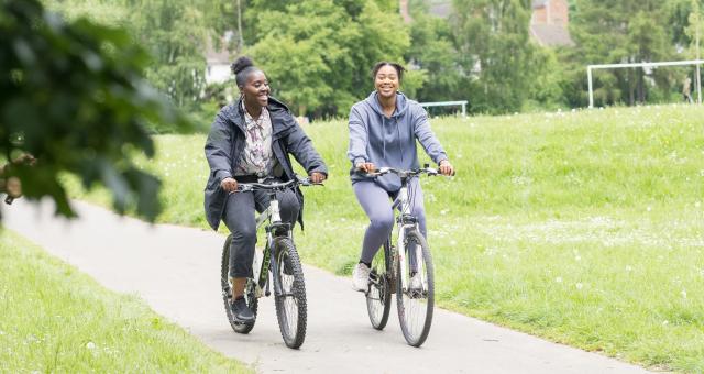 Two women are cycling on a paved path in a park. They are both riding hybrid bikes. One is wearing black jeans, trainers, a patterned shirt and grey coat. The other is wearing a grey tracksuit and white trainers. 