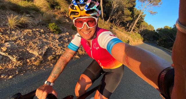 A woman is taking a selfie as she rides along a tarmac road in Mallorca. It’s sunset. She’s wearing cycling kit, including helmet and sunglasses. She’s smiling into the camera