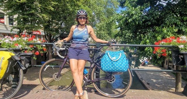 A woman is leaning against a touring bike leaning against a fence on a bridge across a canal. She is wearing shorts and a sleeveless T-shirt, sunglasses and a helmet. There are a lot of trees and flowers in planters in the background.