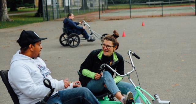 A man and a woman are on adapted cycles in a sports ground. They are both wearing jeans and fleeces