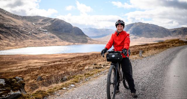 A woman is standing astride a packed off-road bike on a gravel track through mountainous countryside with a lake in the background. She is wearing red, winter cycling jacket, winter gloves and trousers, as well as a helmet. She is smiling at the camera
