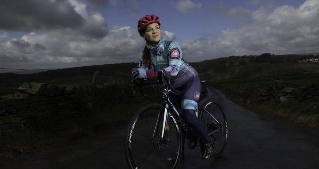 Vicky Mathwin is standing astride her road bike with one foot on the pedal on a country lane. She is wearing full racing cycling kit and a helmet.