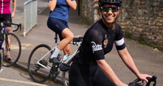 Rachael Burnside is sitting astride a red road bike. She is wearing black cycling shorts and a black and white short-sleeved jersey, as well as a helmet and cycling sunglasses. She’s smiling at the camera. In the background are two more women on road bikes.