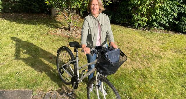 A woman is standing with her hybrid bike. She is wearing blue jeans, trainers and a pale green jacket. Her bike has a basket on the front with her handbag in it. She has blonde hair and is smiling at the camera