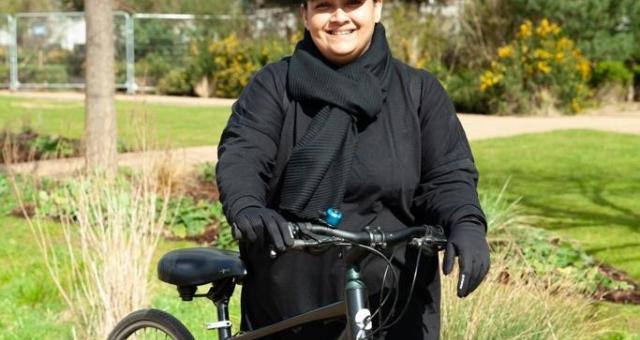 A woman is standing astride holding a bike in a park. She is wearing a black scarf and long black dress, as well as a black cap. Her bike is a dark metallic green hybrid. She is smiling at the camera. Photo: Sonny Malhotra