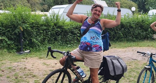 ALT TEXT: Sara Ramsey is sitting astride a black Trek touring bike. She is wearing shorts and a T-shirt. She has her arms up in ‘strong man’ pose and she’s smiling at the camera.