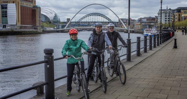 Three people are standing astride their bikes next to the River Tyne. The BALTIC and Sage and the Tyne bridges can be seen in the background