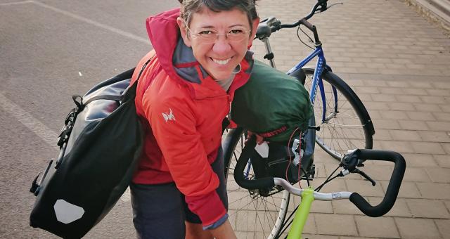 Susanna Thornton is looking up at the camera while holding onto a bike. She has two packed panniers slung across her shoulders. She is wearing shorts and a jacket and glasses; she also has sunglasses on her head. She is smiling.