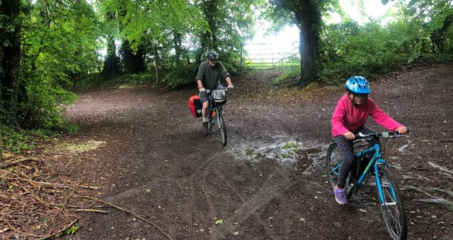 Two people are cycling through a muddy forest: a young girl on a blue mountain bike is in front and she's followed by a man on a flat-bar touring bike with packed pannier and bar bag.