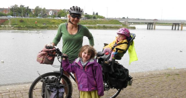 A woman in a green top is standing holding a bike. She has two little girls with her. One is standing in front of the bike and is wearing a purple coat and yellow dress, the other is in a child seat on the back of the bike