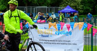 A man stands stationary with a bicycle in a park in front of a banner which says the Big Bike Revival.