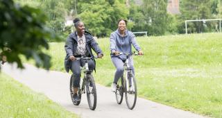Two women are cycling on a paved path in a park. They are both riding hybrid bikes. One is wearing black jeans, trainers, a patterned shirt and grey coat. The other is wearing a grey tracksuit and white trainers. 