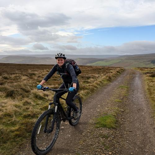 A cyclist wearing a bright green jacket and a helmet rides a mountain bike along a rugged trail through a wild and open moorland. The cloudy sky and expansive landscape create a sense of adventure and connection with nature, highlighting the joy and resilience of off-road cycling.