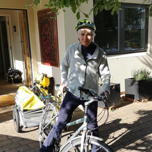 A woman is standing astride a grey hybrid bike with a packed trailer attached to it. She is wearing normal clothes: blue trousers and shoes and a grey fleece, as well as a cycle helmet.