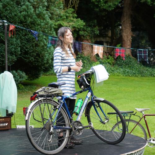 A woman is standing on a makeshift stage. She has a Dawes touring bike with her; an open notebook is balanced on the bike’s handlebars. She is wearing a blue and white striped top, black three-quarter-length trousers and cycling shoes. She is smiling.