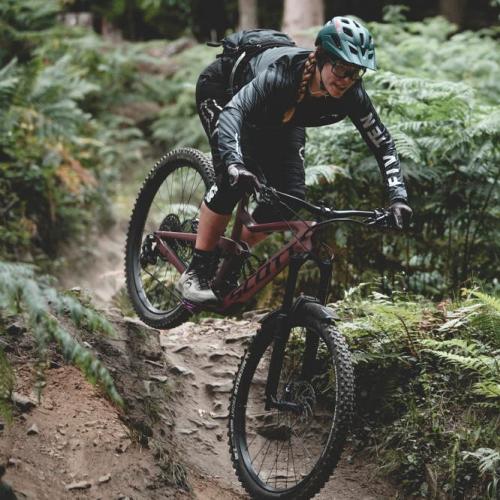 A woman is cycling downhill through a forest. She is riding a Scott mountain bike. She is wearing black three-quarter-length cycling leggings and a black jacket, as well as a green helmet. Credit: Roo Fowler