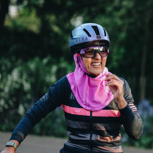 A woman in cycling gear is smiling at the camera. She is wearing a blue cycling helmet with a Rapha cap underneath. She has on cycling sunglasses, a stripey Rapha cycling jersey and a bright pink headscarf