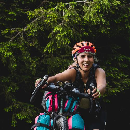 A woman is pushing her bike uphill. Only the bike’s handlebars and top of the front wheel are in shot, along with packed bikepacking bags. She is wearing an orange helmet and a Rapha cap